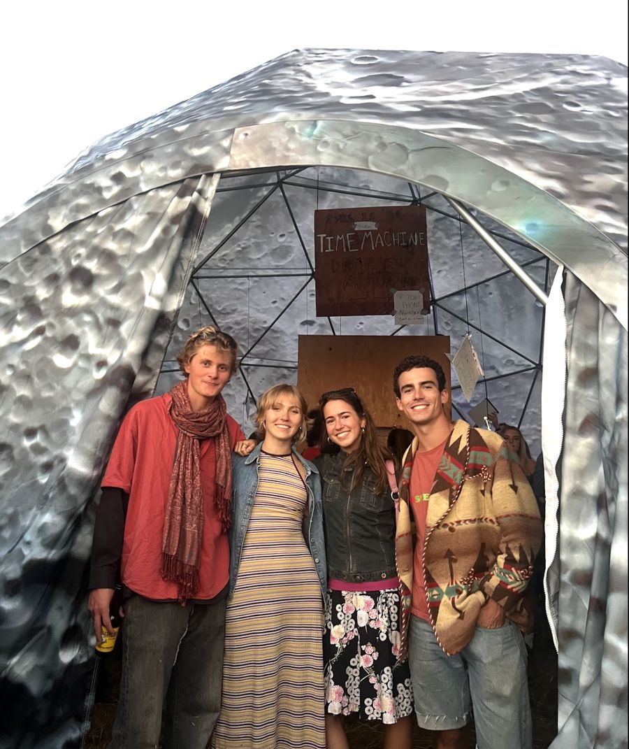 Four people standing at the entrance of a geodesic dome painted like the moon, with a hand-lettered sign reading THIS IS A TIME MACHINE, DROP YOUR MEMORY