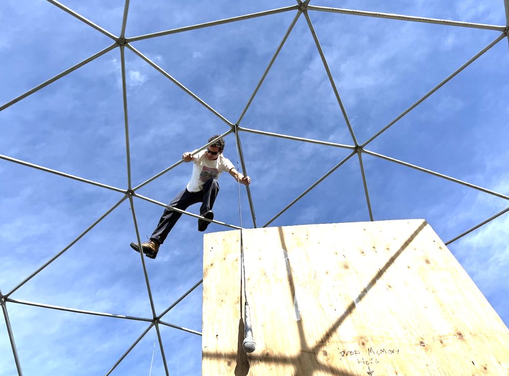 G climbing the exposed steel frame of the geodesic dome above a plywood panel with a hanging microphone