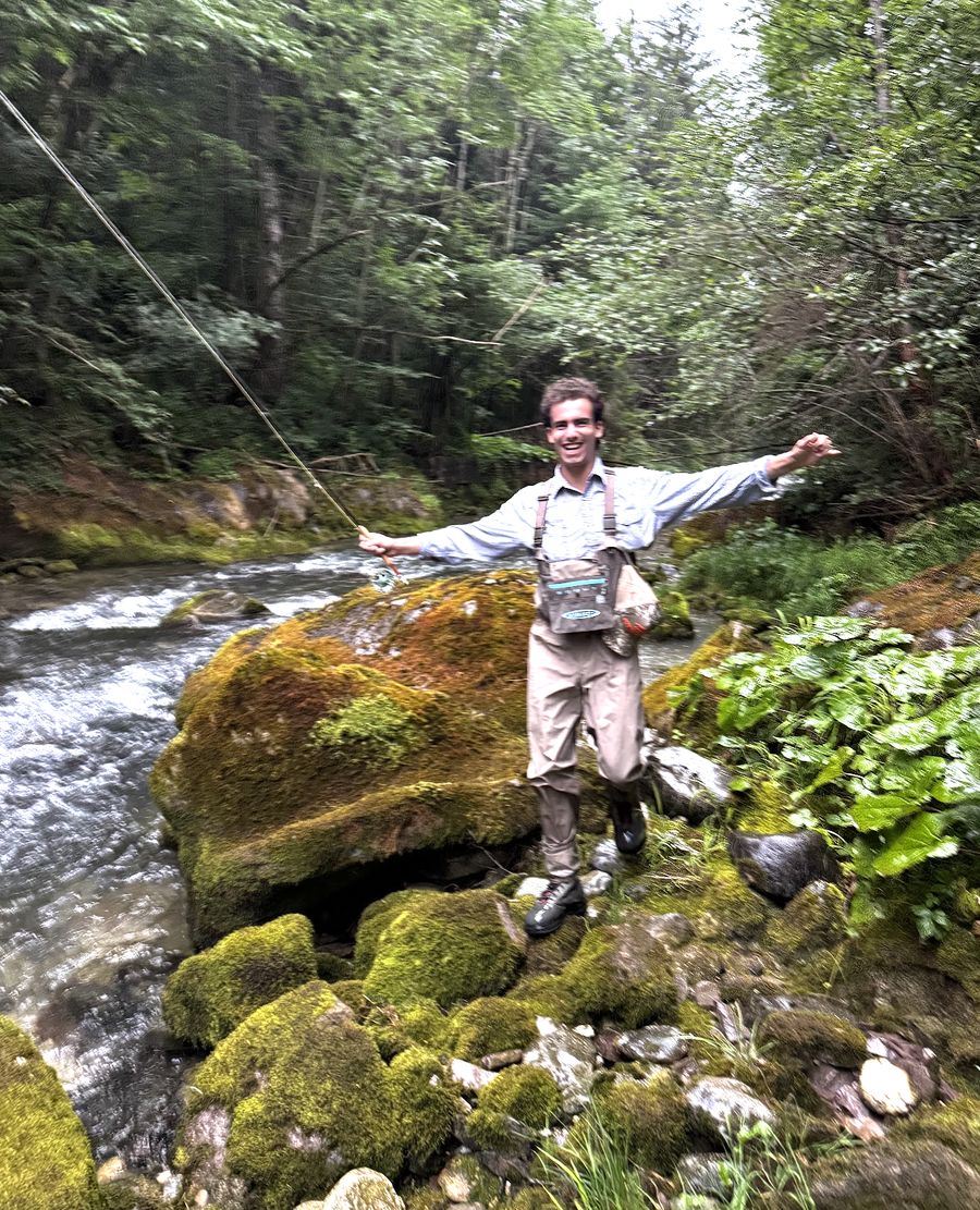 G in waders on mossy boulders beside a fast alpine river, fly rod bent, arms out